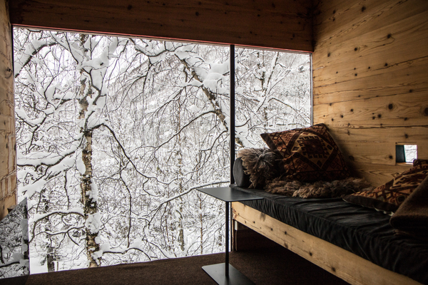 Interior view from a cozy wooden cabin showing a large window framing a snowy birch forest, with a built-in daybed covered in pillows.