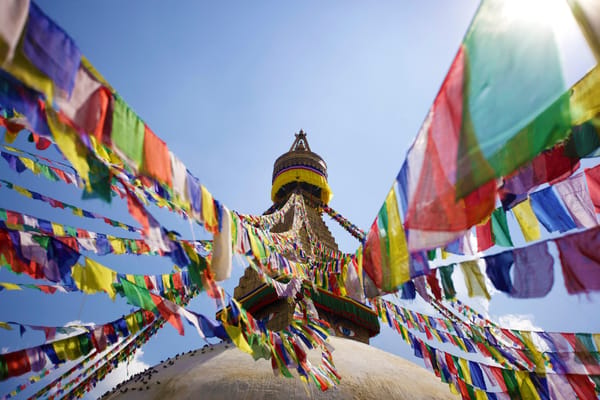 Looking up from the base of a massive white stupa, countless colorful prayer flags radiate outward from its golden spire, which features painted Buddha eyes, against a clear blue sky.