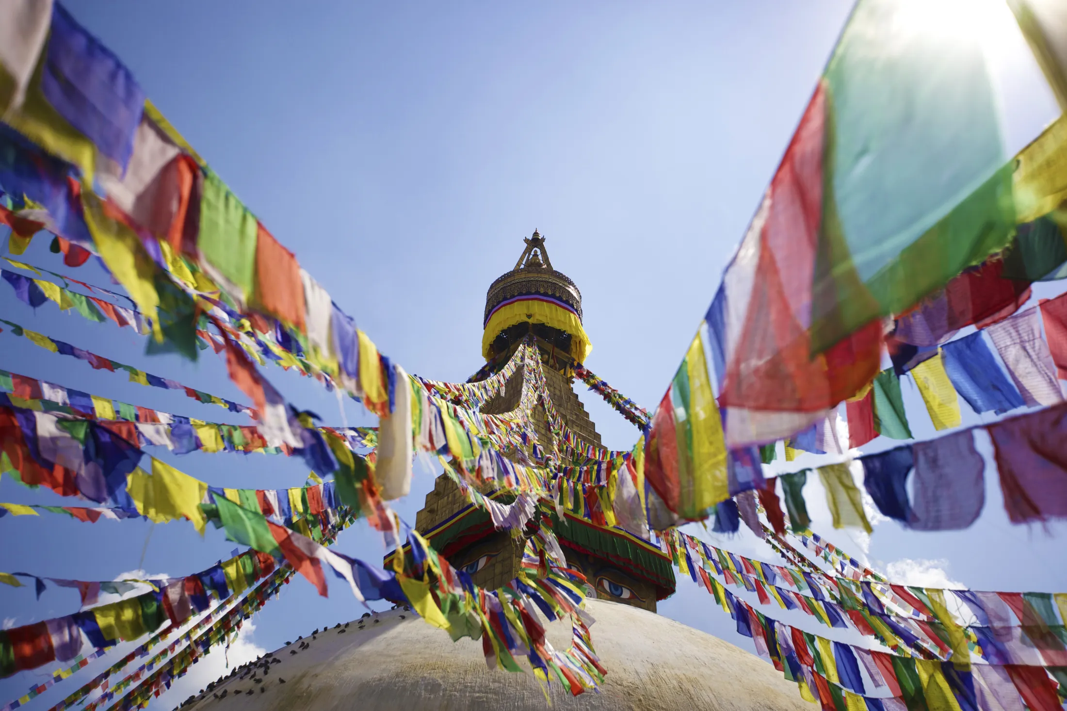 English: A low-angle shot looking up at the Boudhanath Stupa, with colorful prayer flags radiating outwards against a clear blue sky.