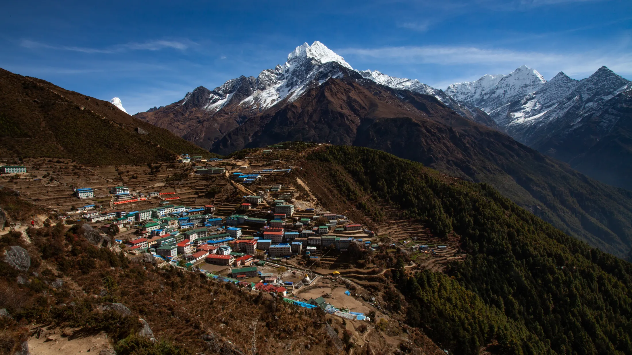 A vibrant hillside village with colorful roofs sits nestled on a mountain slope, with a massive, snow-capped peak rising majestically into the blue sky in the background.