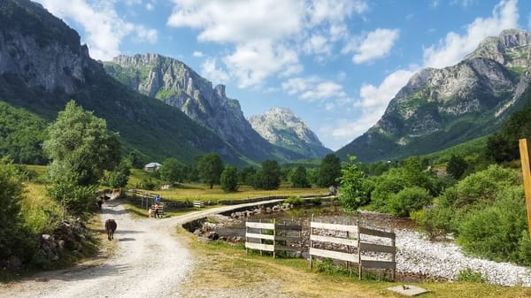 A dirt path through a lush green valley surrounded by majestic mountains under a blue sky.