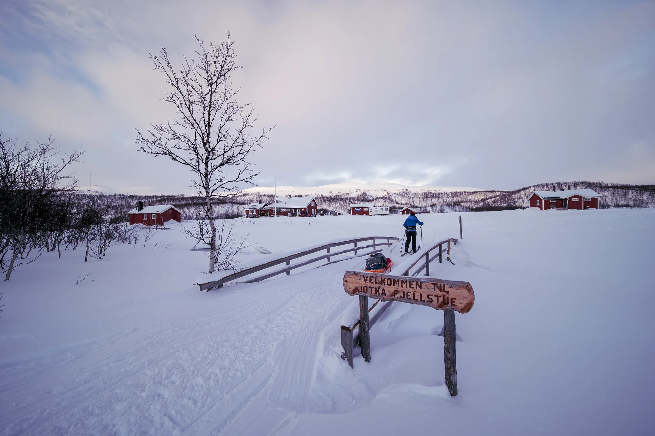 A person on a small wooden bridge crossing a snowy landscape with red cabins in the background under a cloudy sky.
