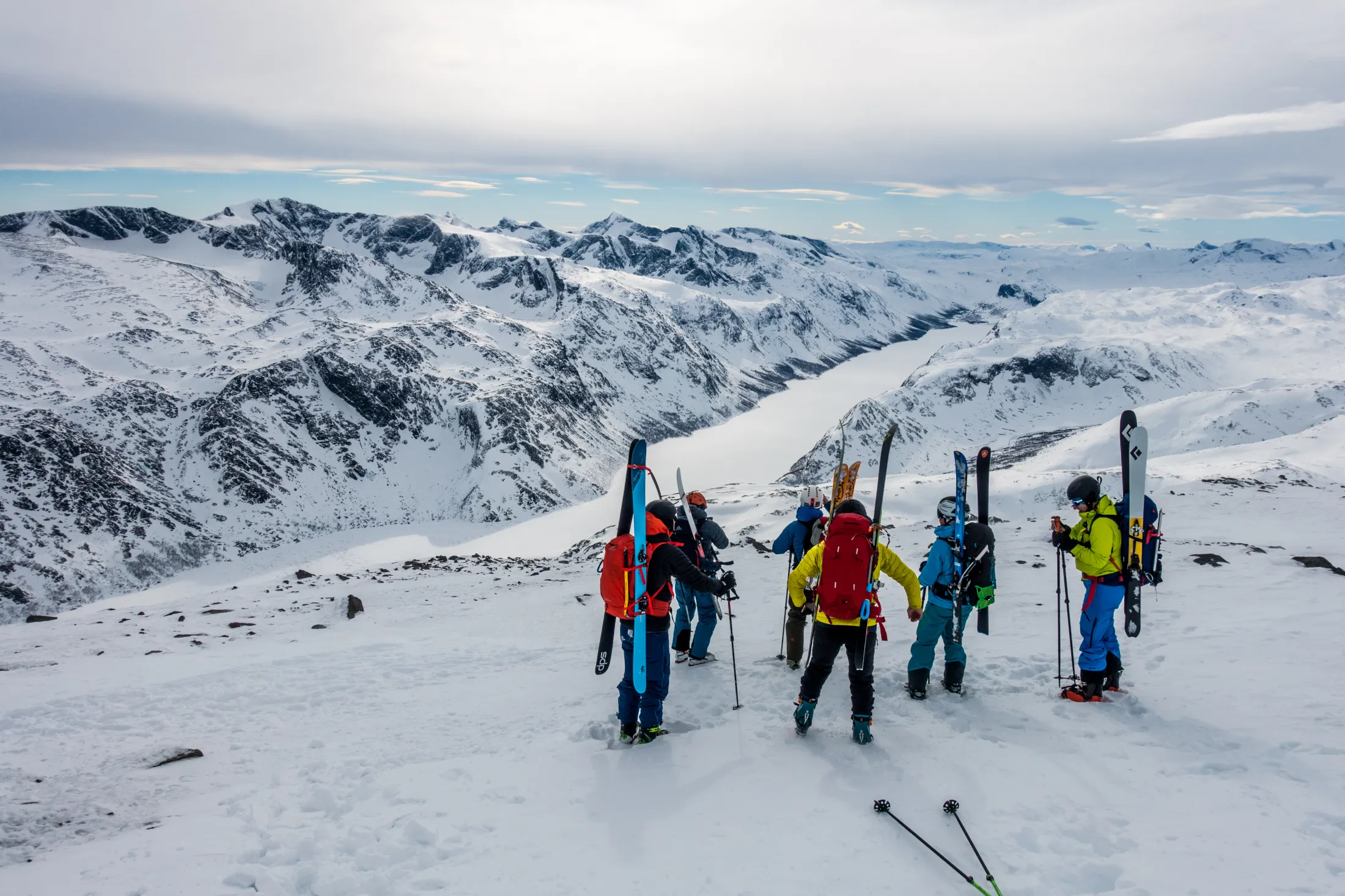 A group of skiers in colorful winter gear stand on a snowy mountain ridge, overlooking a vast snow-covered valley and rugged mountain peaks under an overcast sky.