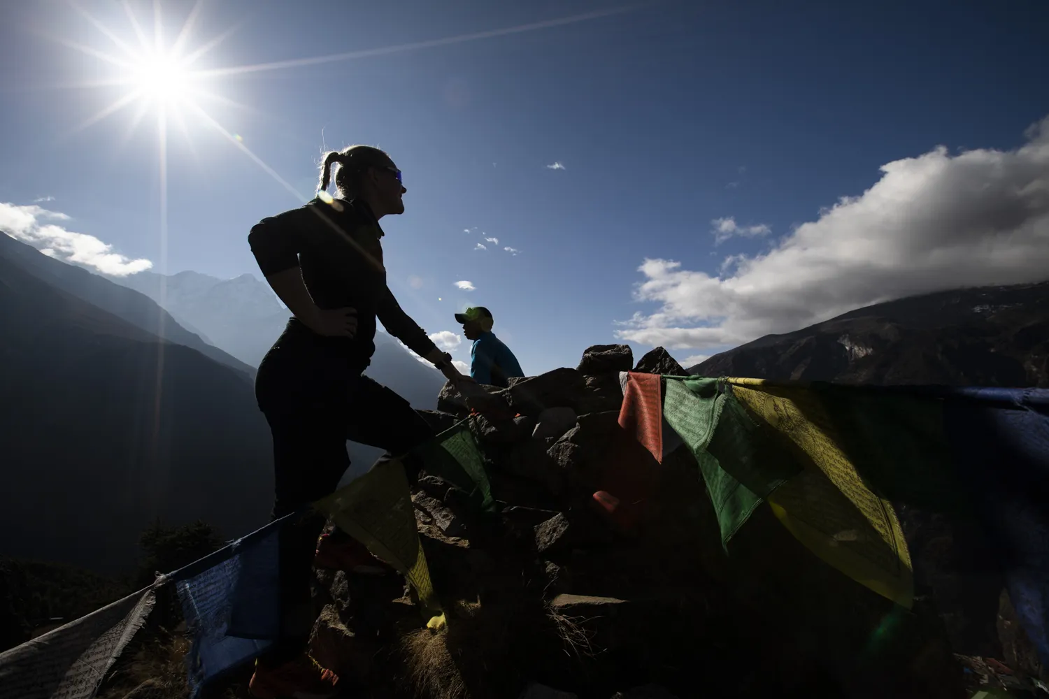 A low-angle silhouette captures a female hiker standing on a rocky ridge beside colorful Tibetan prayer flags, looking out at mountain ranges under a bright, sun-drenched blue sky.