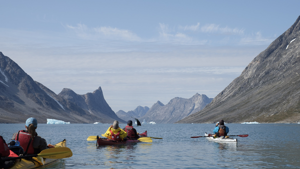 Several kayakers in a narrow fjord watch a whale tail breaking the surface of the water in the distance, surrounded by tall, steep mountains under a partly cloudy sky.