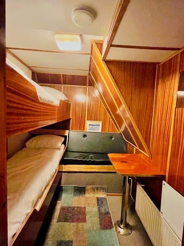 A narrow boat cabin interior featuring wooden paneling, bunk beds on the left, a small seating area with a table in the center, and a patterned rug on the floor.
