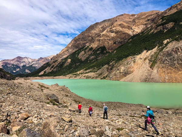 Four hikers approach a turquoise glacial lake surrounded by rocky mountains.