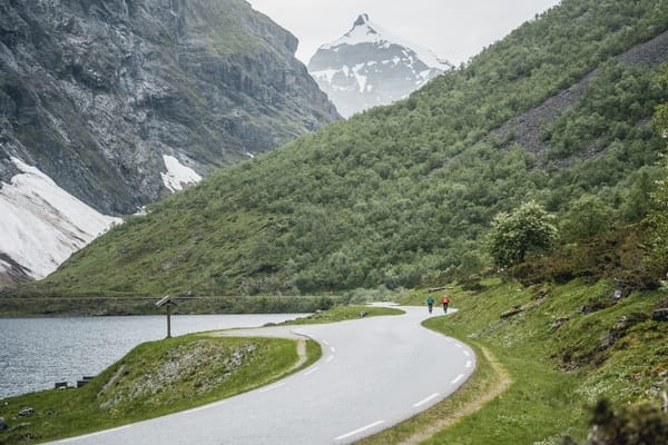 A winding mountain road near a lake surrounded by steep, snow-dusted peaks in Norway.