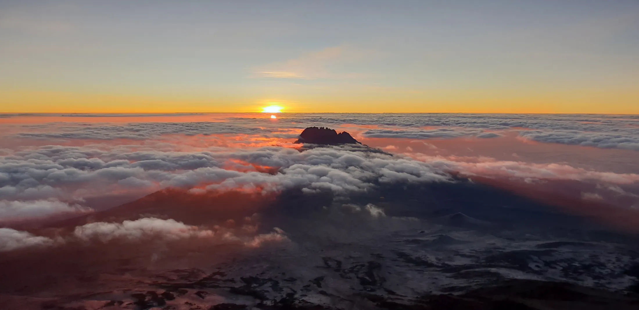The sun rises over a sea of clouds surrounding the dark silhouette of a mountain peak.