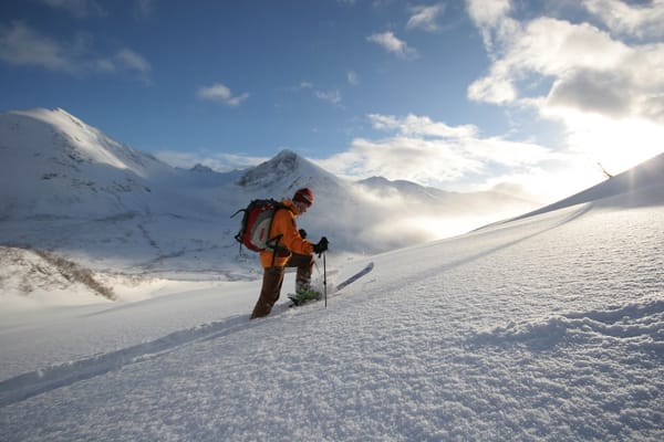 A ski tourer climbing a snowy slope against a backdrop of sunlit mountain peaks and a blue sky.