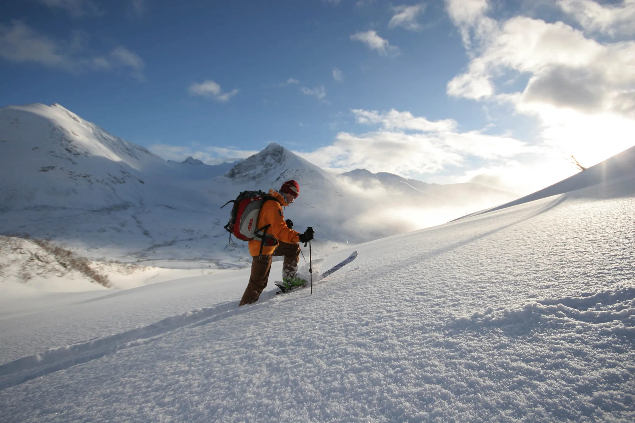 A ski tourer climbing a snowy slope against a backdrop of sunlit mountain peaks and a blue sky.