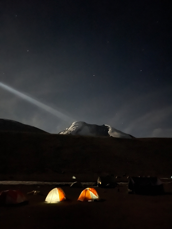 Two glowing orange tents illuminate a dark base camp at night, with a faint beam of light cutting through the starry sky above a distant snow-covered mountain peak.