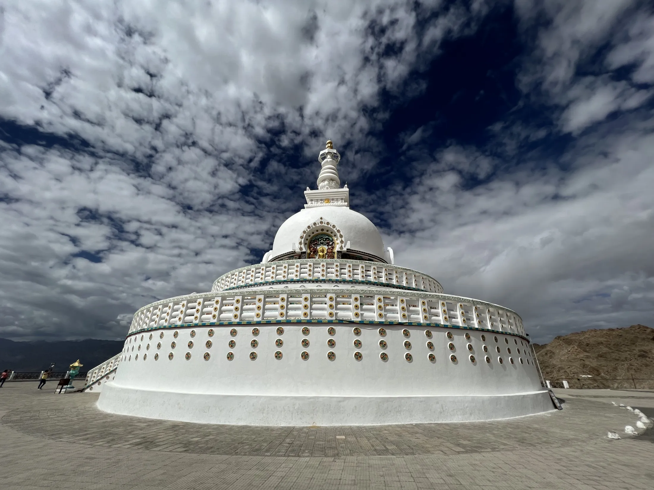 A large, white Buddhist stupa decorated with gold and circular reliefs stands dramatically against a striking blue sky filled with textured white clouds.
