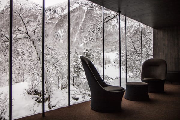 Interior view through a large glass window showing a snowy landscape outside, with dark armchairs and a stool placed inside.