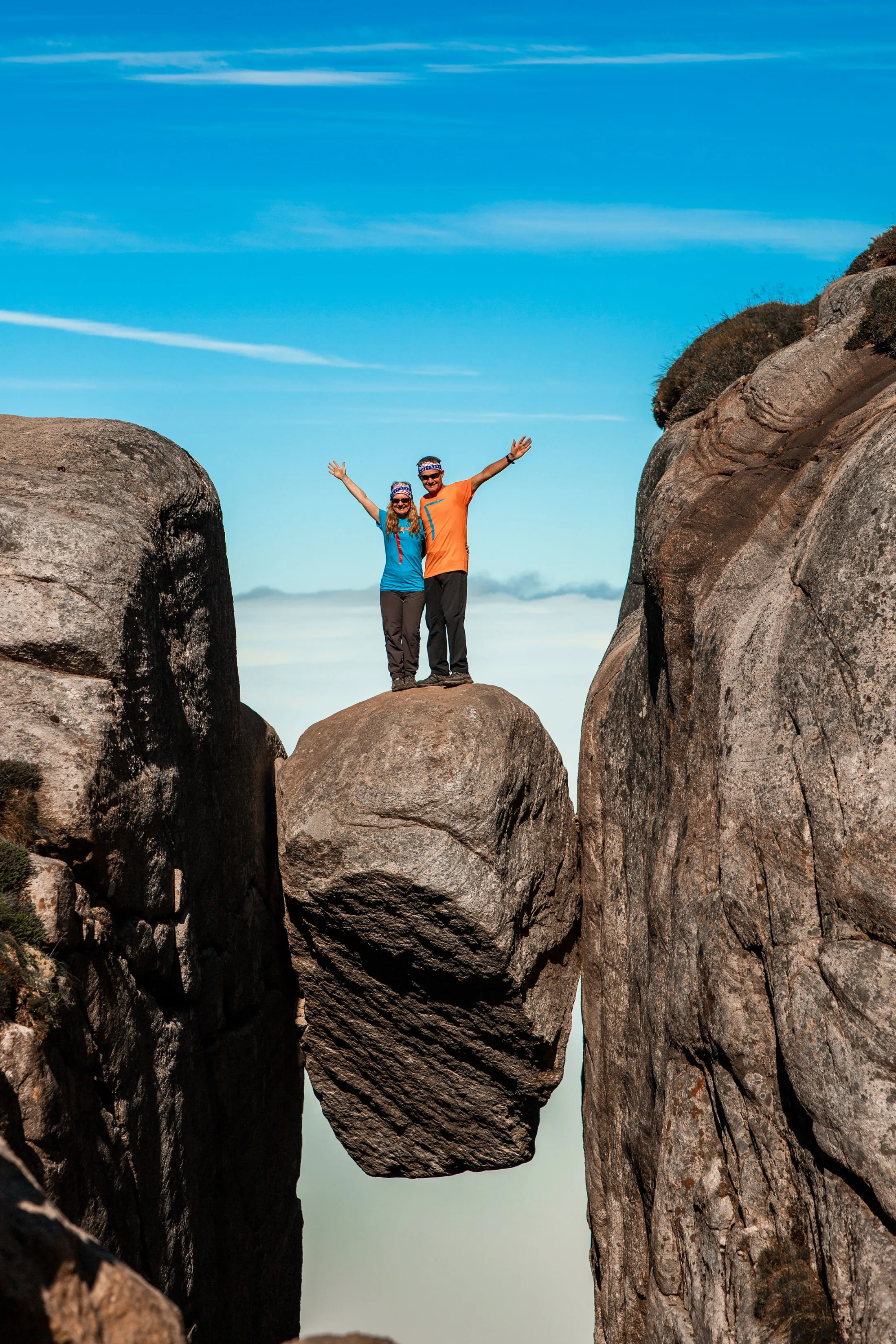 Two people standing on Kjeragbolten, a boulder wedged in a mountain crevasse in Norway, with arms raised in celebration against a blue sky.