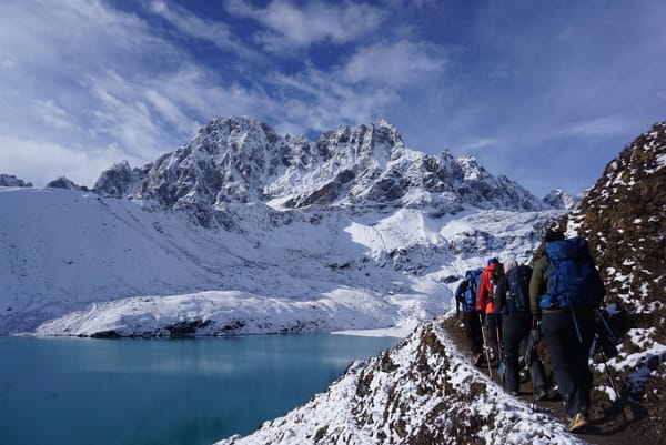 A line of hikers with backpacks walks along a snowy trail beside a vibrant turquoise alpine lake, heading toward massive snow-capped mountains under a blue sky.