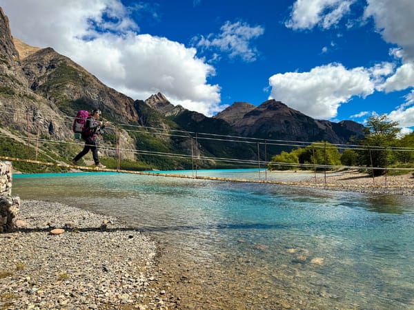 A hiker with a backpack crosses a suspension bridge over a turquoise river in a mountain landscape.