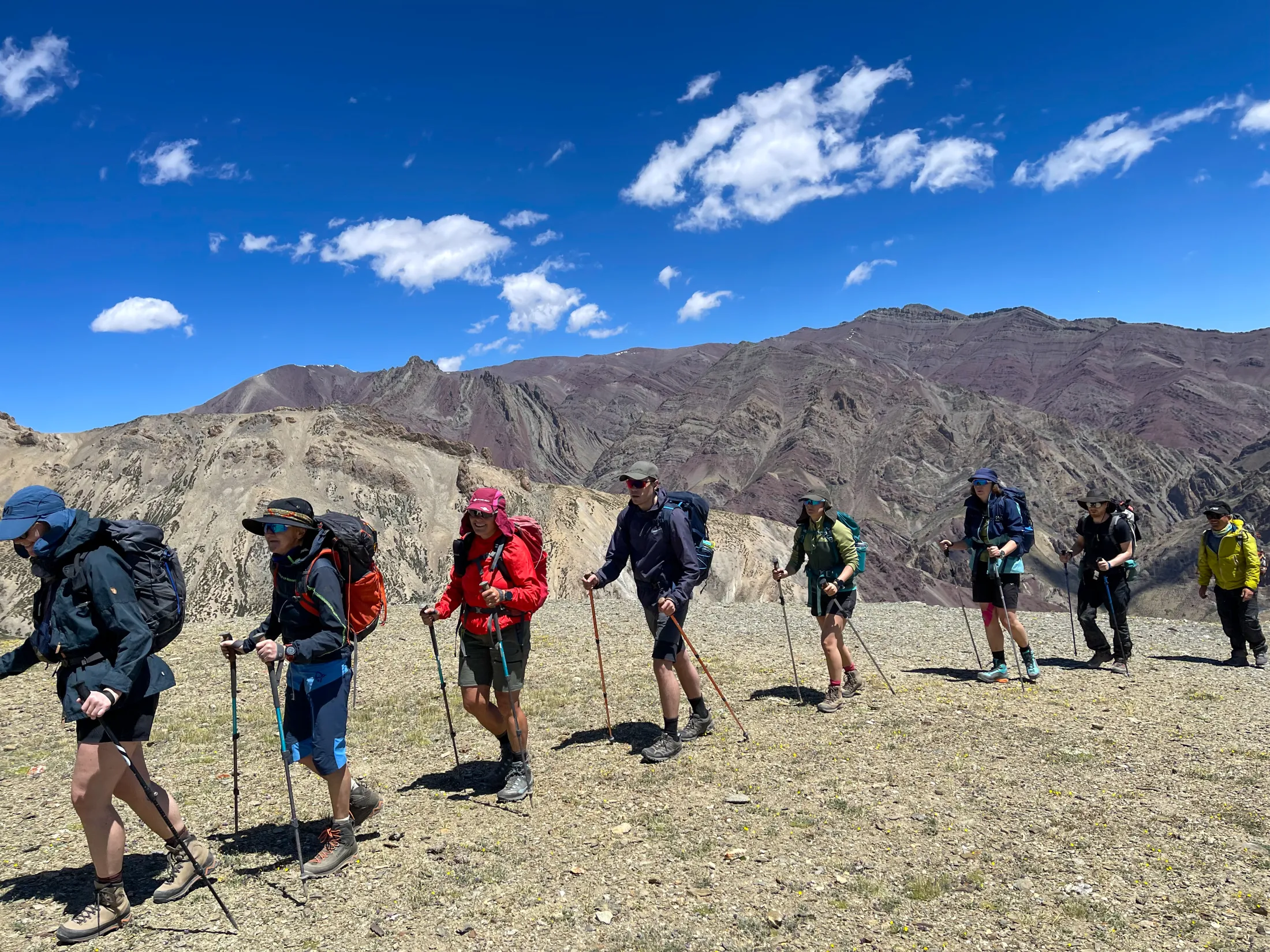 A group of hikers with backpacks and trekking poles walks in a line across a dry, gravelly mountain ridge under a bright blue sky with scattered clouds.