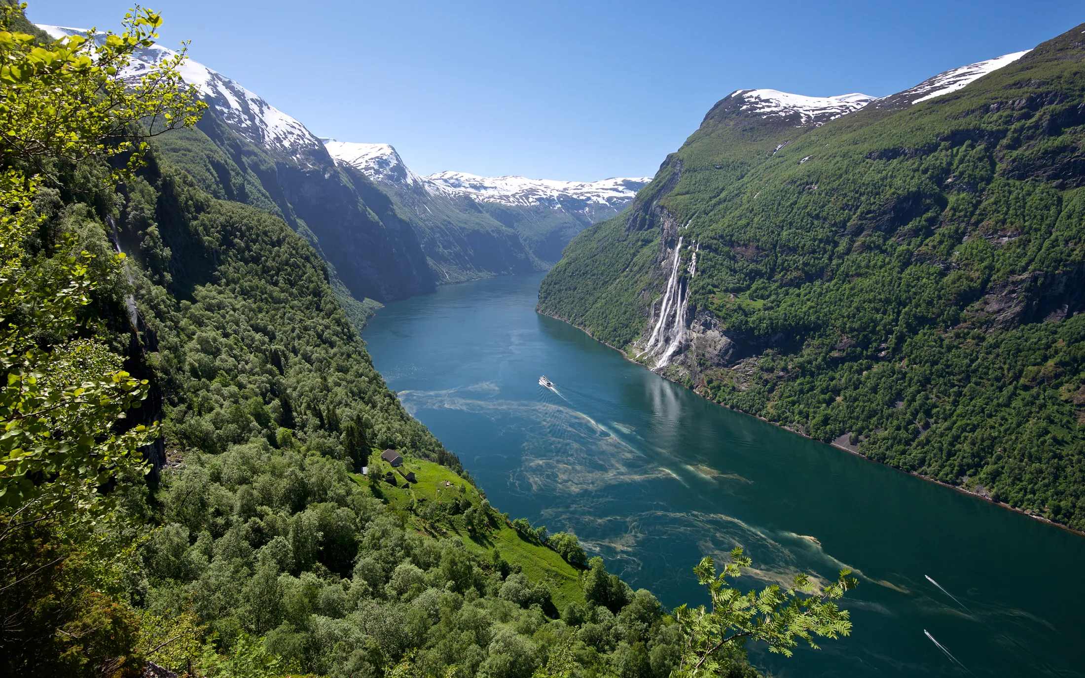 The Geirangerfjord in Norway, surrounded by steep, green mountains and snow-capped peaks.