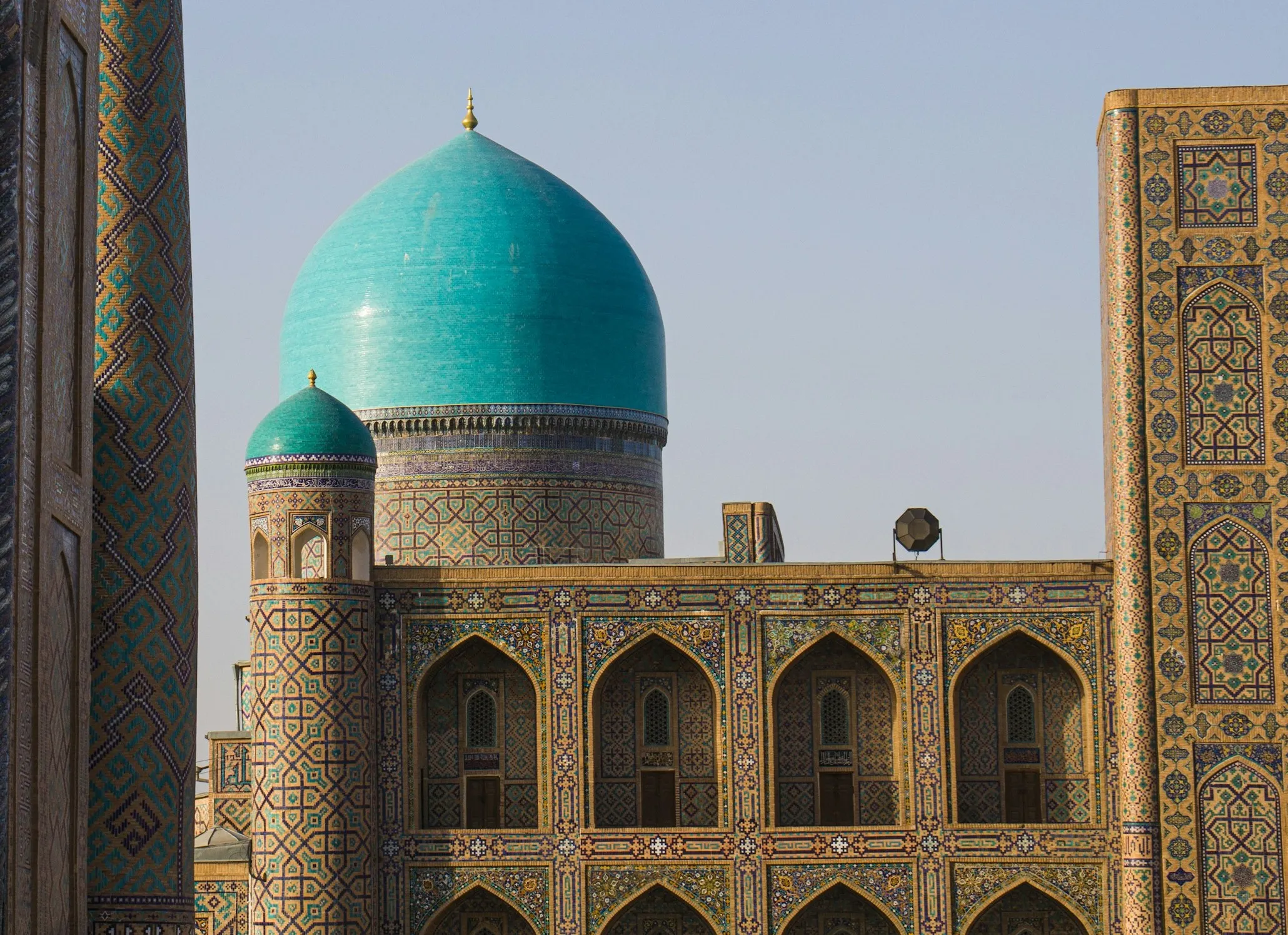 A grand turquoise dome rises above an intricately tiled madrasah facade featuring arched alcoves and patterned geometric designs.