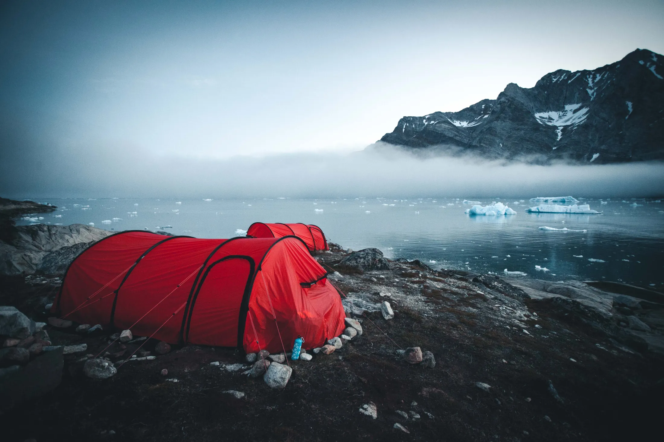 Two red tunnel tents are pitched on a rocky shoreline, overlooking an ocean filled with drift ice and low-hanging fog in front of dark mountains.