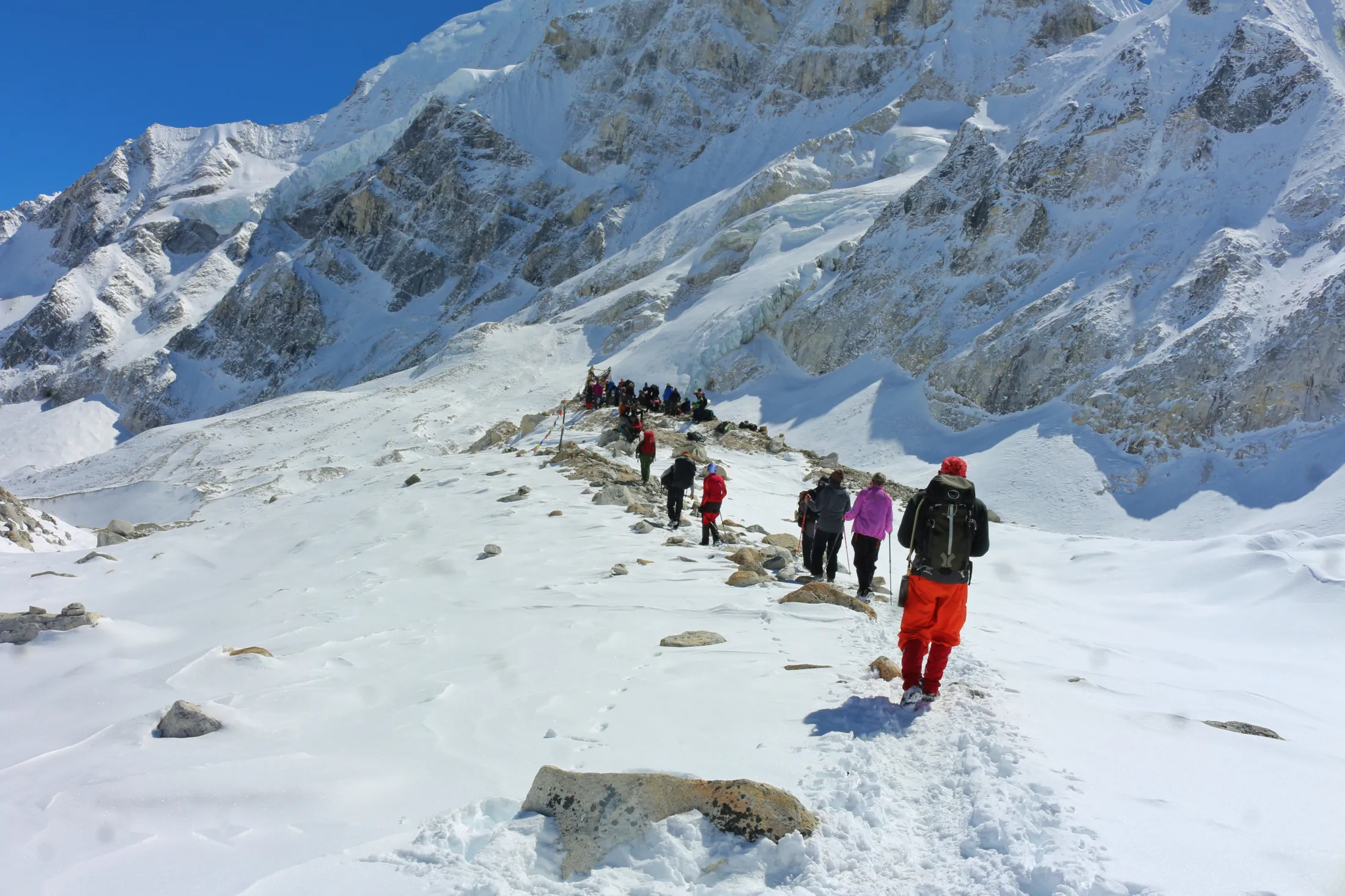 A group of mountaineers treks along a snow-covered ridge leading toward a massive, icy mountain wall under a clear blue sky.
