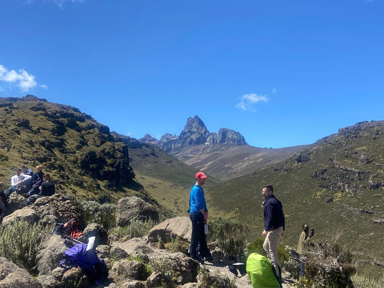 Hikers rest on a rocky, grassy ridge with a clear view down a mountain valley toward the rugged, distant peaks of Mount Kenya under a bright blue sky.