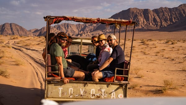 A group of smiling tourists sitting in the back of an open pickup truck driving through a desert landscape.
