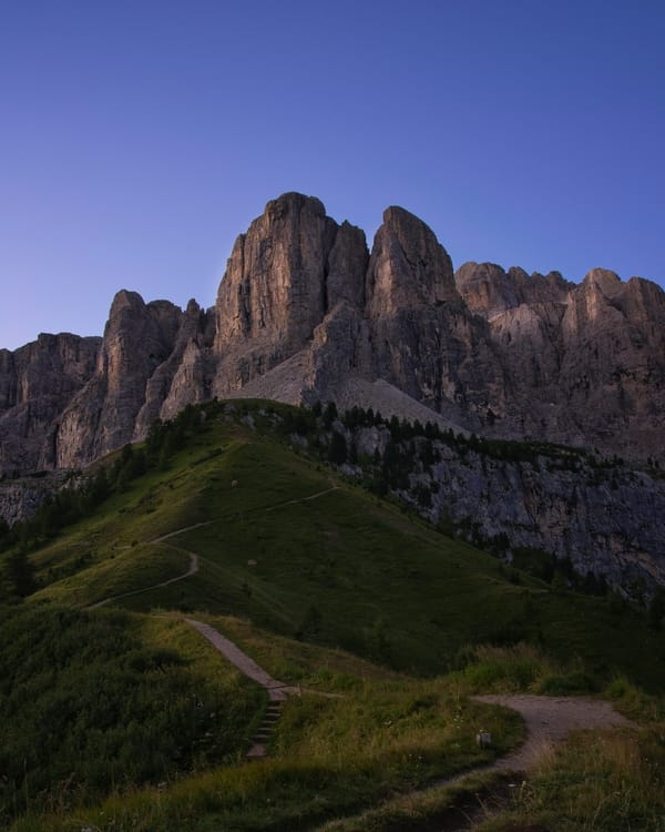 A hiking trail winding up a grassy hill toward sharp mountain peaks under a clear blue sky at dusk.