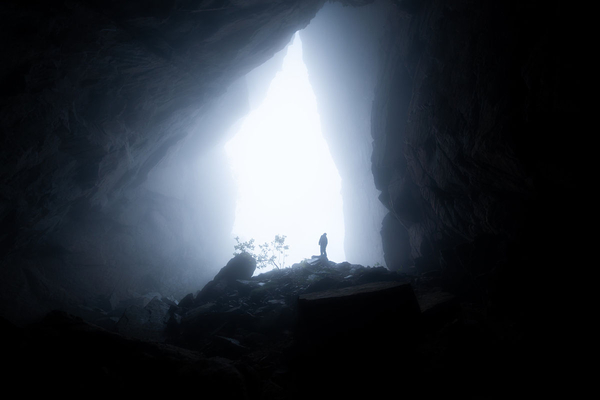 A person stands silhouetted at the entrance of a dark cave.