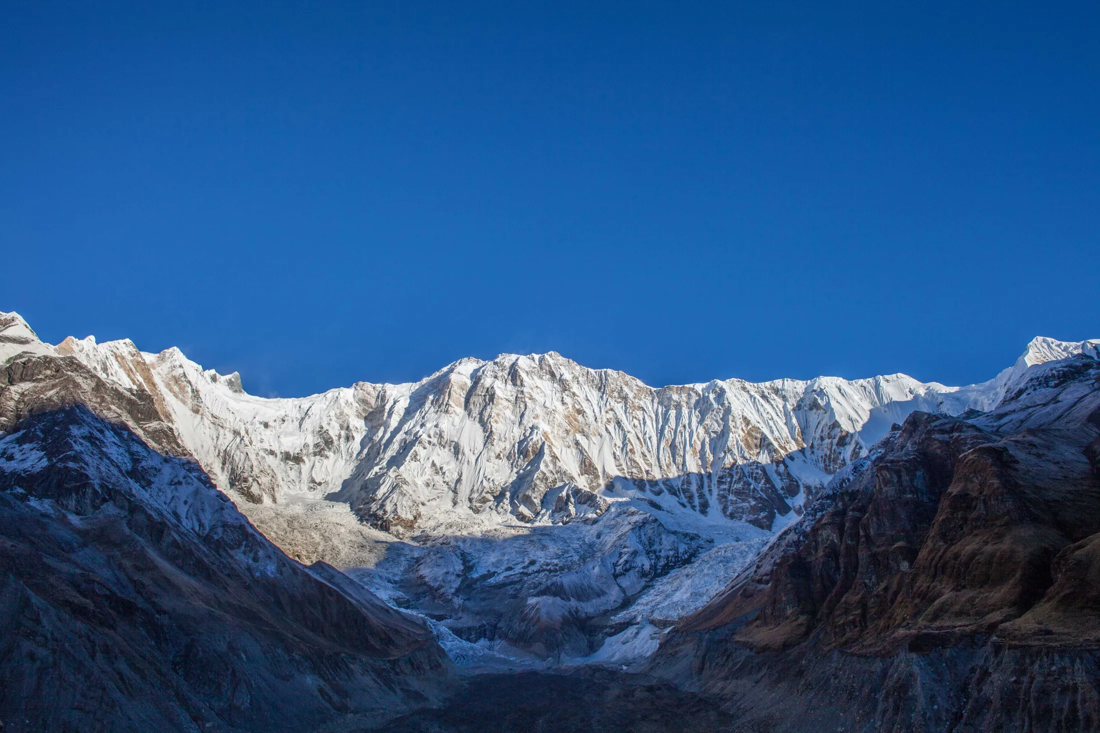 A colossal, snow-dusted mountain wall with intricate, ice-filled ridges towers over a deep, dark valley under a vast, clear blue sky.