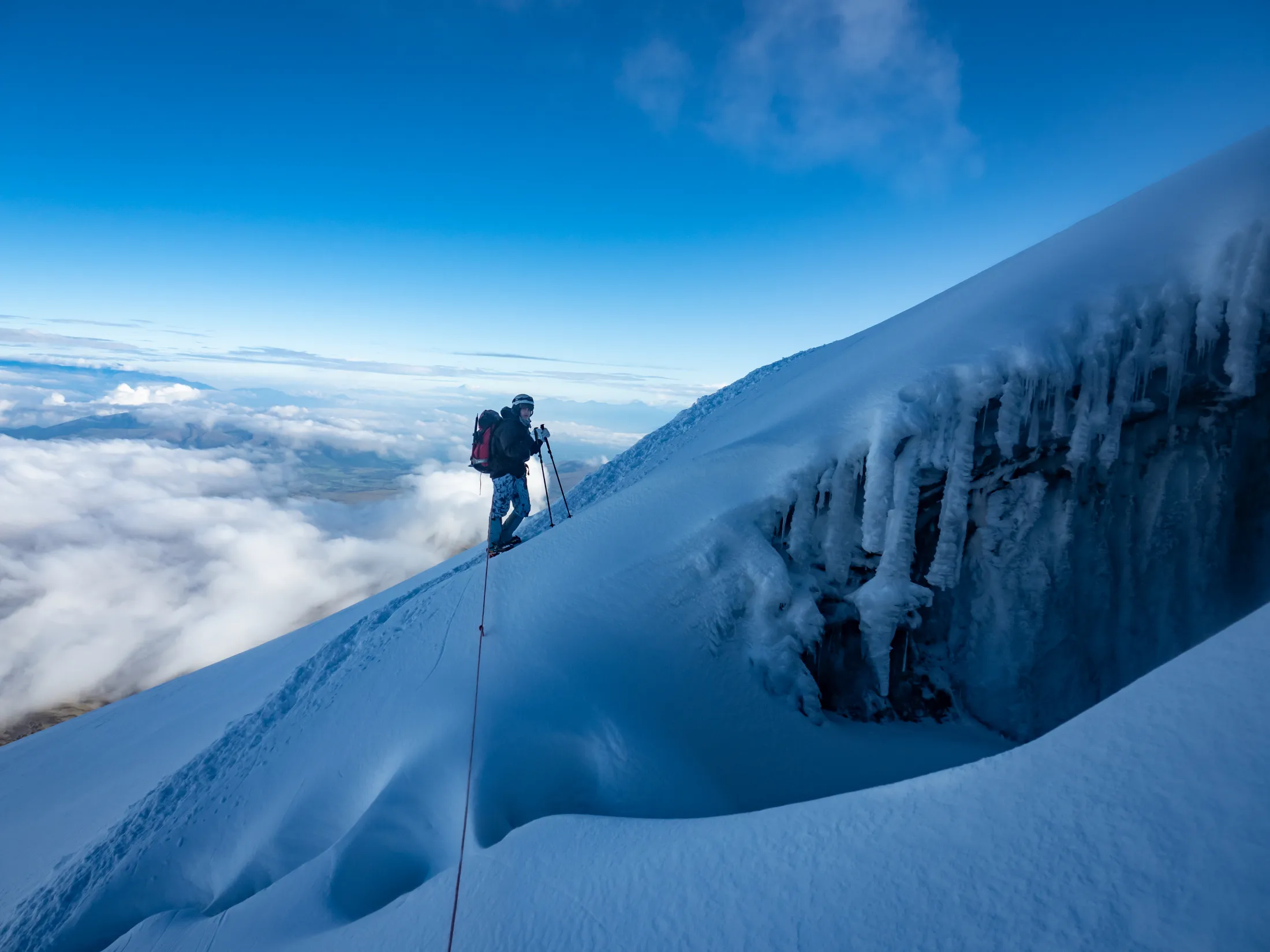 A lone mountaineer walks along a snow ridge above a sea of clouds under a clear blue sky.