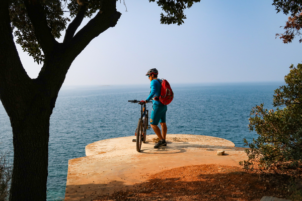 A cyclist with a red backpack stands on a concrete platform overlooking the sea.