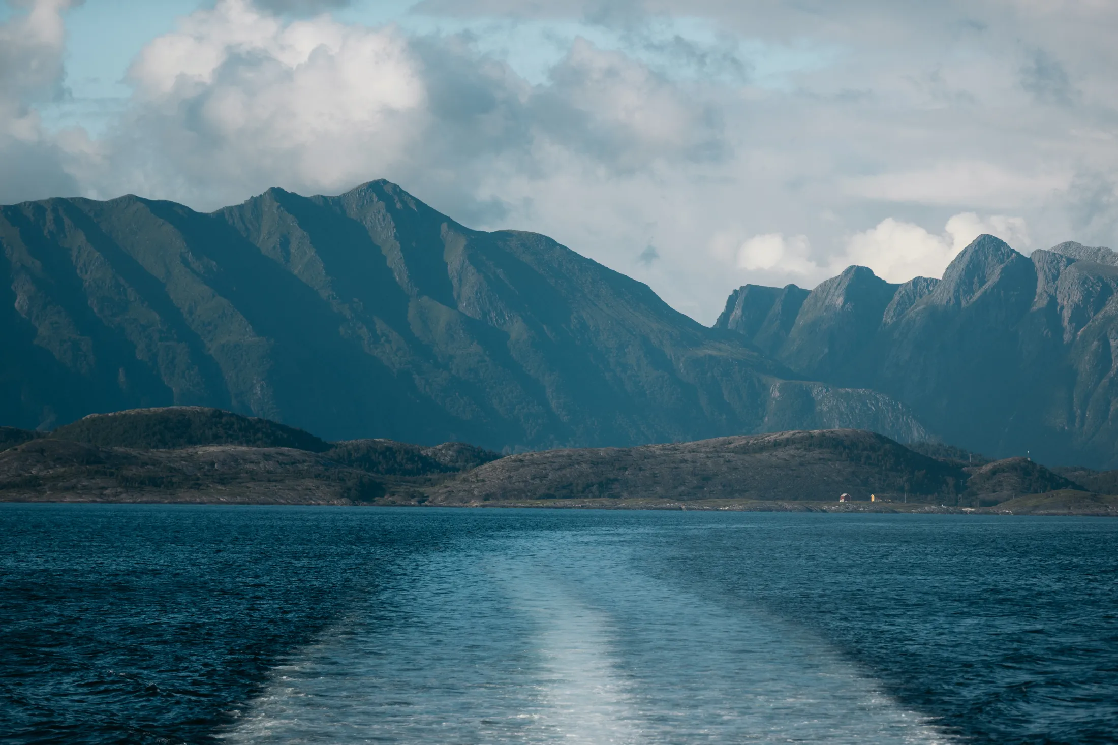 A wake left in the sea by a boat traveling towards mountains under a cloudy sky.