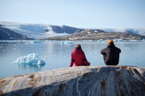 Two people sit on a rocky shore looking out over a blue fjord filled with light blue icebergs, with a large glacier and snow-covered mountains in the background.