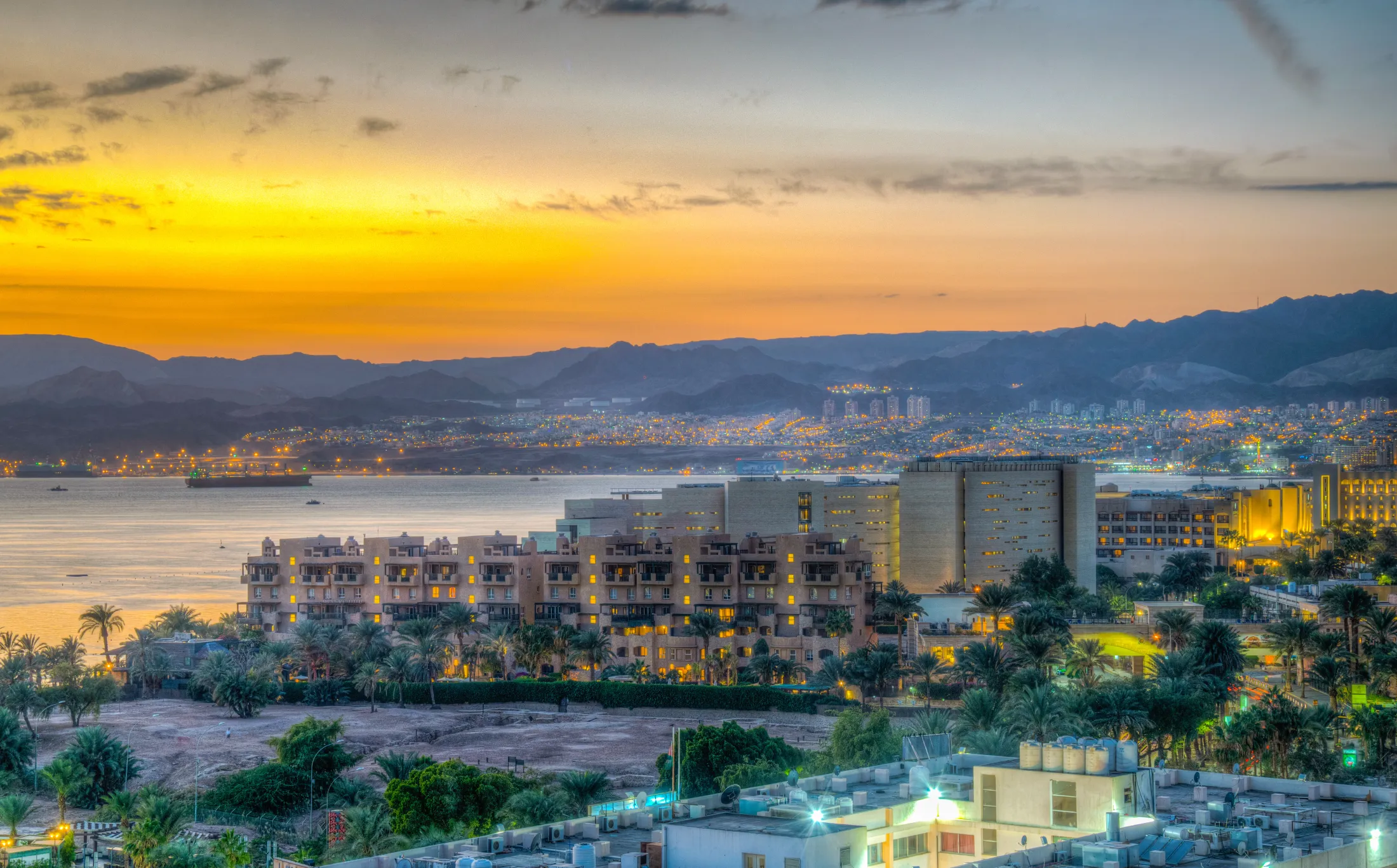 An elevated view of an illuminated coastal city along the water during a golden sunset with mountains in the background.