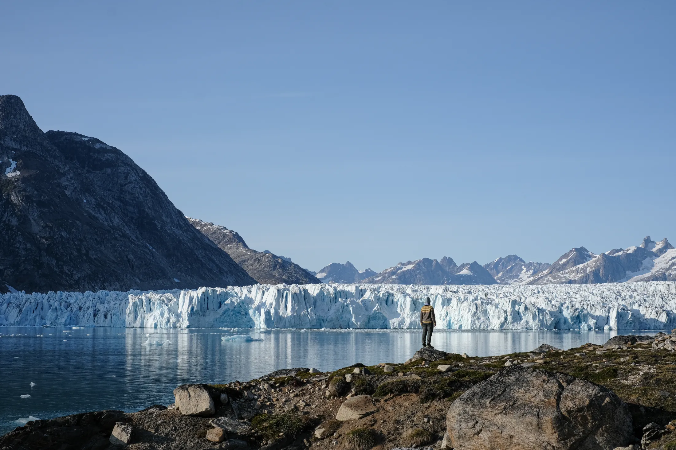 A person stands on a rocky ridge looking out over a massive, light blue glacier wall stretching across a calm fjord under a clear blue sky.