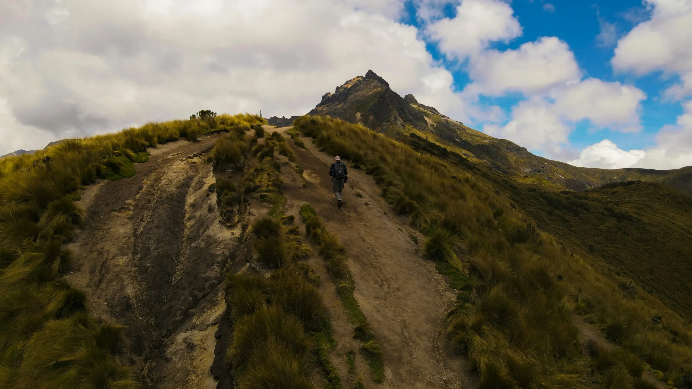A hiker ascends a steep, grassy mountain ridge under a partly cloudy sky.