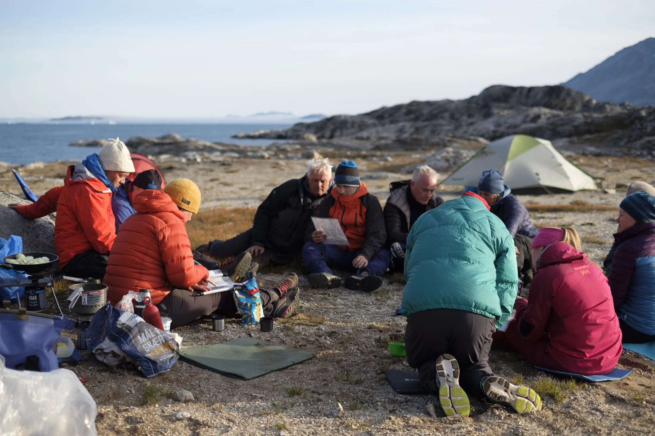 A group of hikers in warm clothing sits in a circle on the ground at a campsite studying maps, with the ocean and rocky terrain in the background.