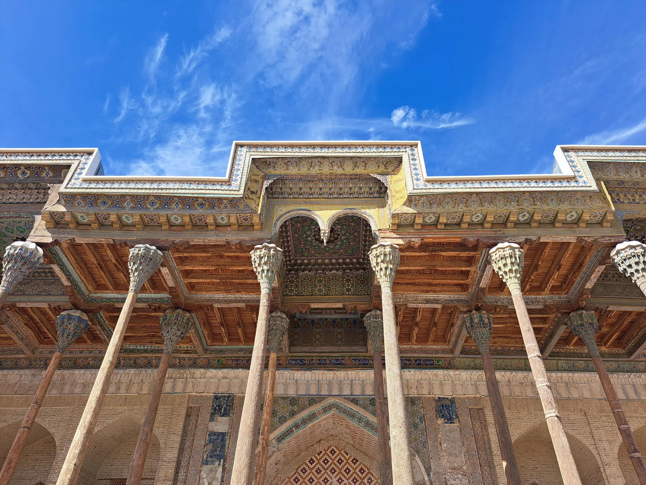 A low-angle view shows an ornate wooden ceiling and intricately carved pillars supporting the grand entrance of a historic building under a bright blue sky.