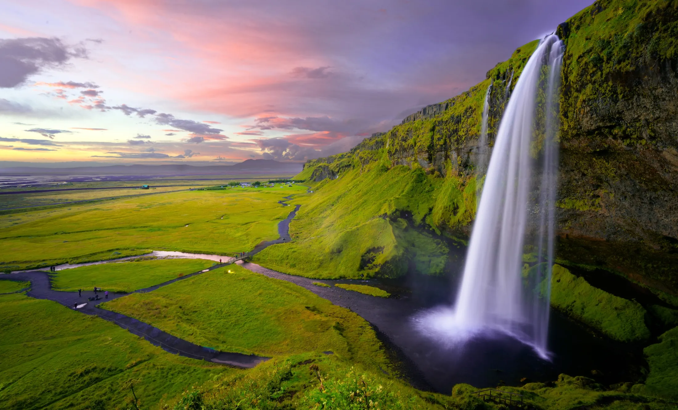 A powerful waterfall cascades down a lush green cliff into a pool next to an open plain under a colorful evening sky.