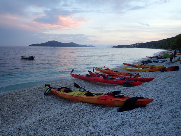 Several red and yellow sea kayaks line a pebble beach at dusk, with a calm sea, a small motorized boat, and distant islands under a pink-tinted sky.