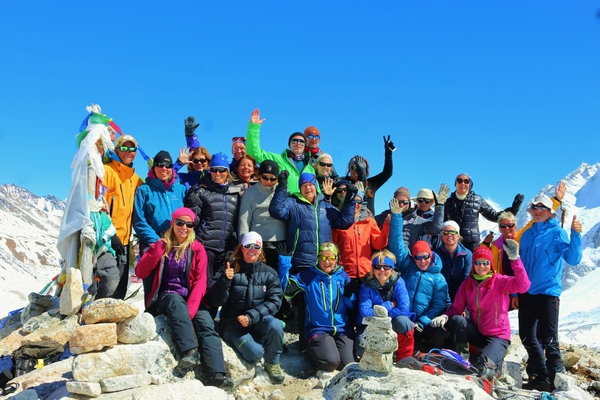 A large group of cheerful hikers in colorful winter jackets and sunglasses poses together on a snowy mountain summit under a brilliant blue sky.
