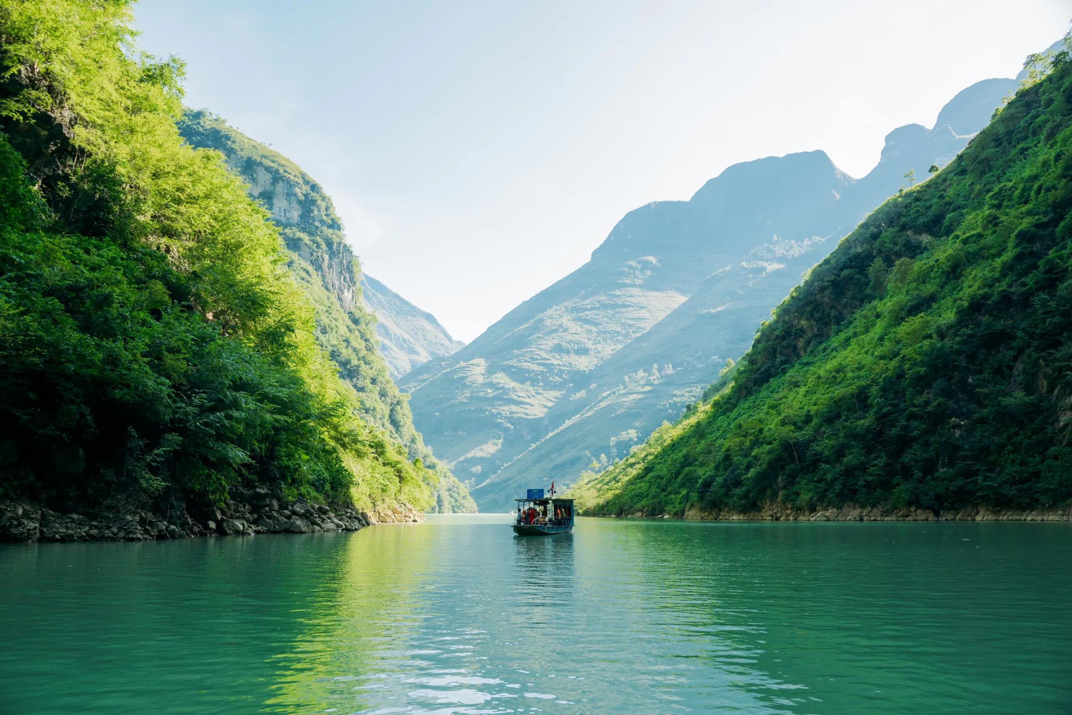 A small boat on a turquoise river flowing between steep, lush green mountains under a bright sky.
