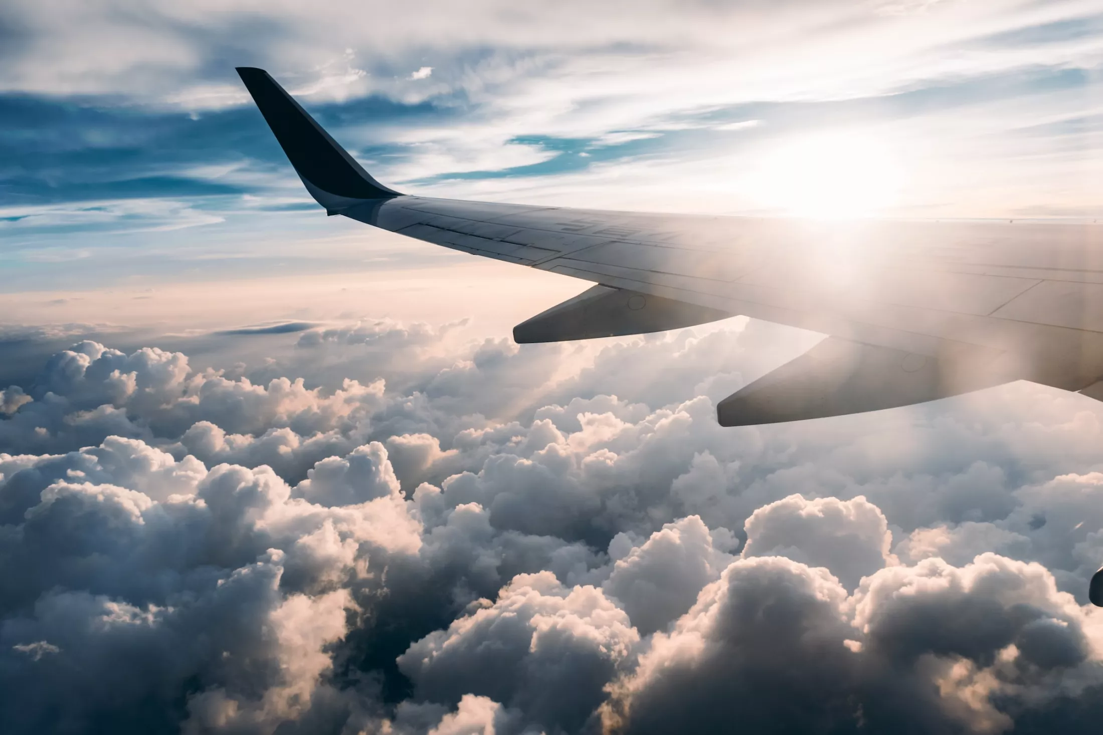 An airplane wing stretches across the sky high above a dense layer of fluffy white clouds with the bright sun creating a brilliant lens flare.