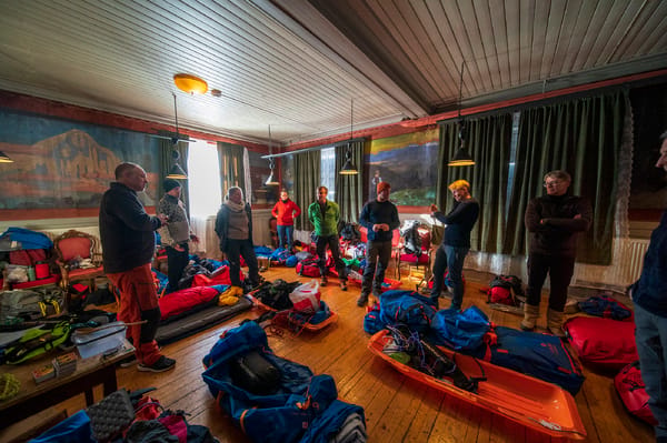 A group of people standing in a large wooden room filled with expedition gear, sleeping bags, and pulks while listening to an instructor.