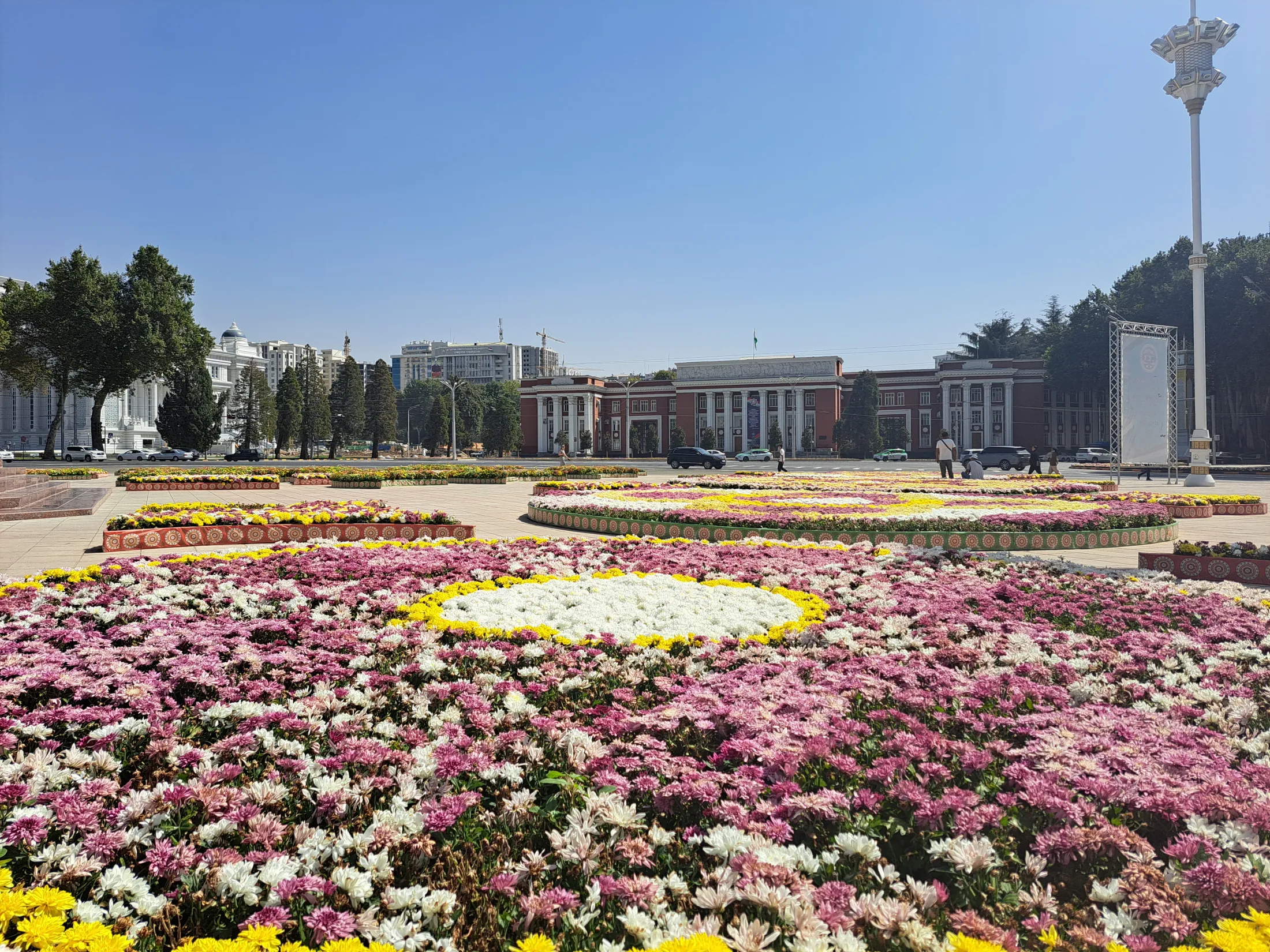 Large, patterned circular flowerbeds filled with pink, white, and yellow blossoms decorate a wide paved public square in front of a grand government building under a clear blue sky.