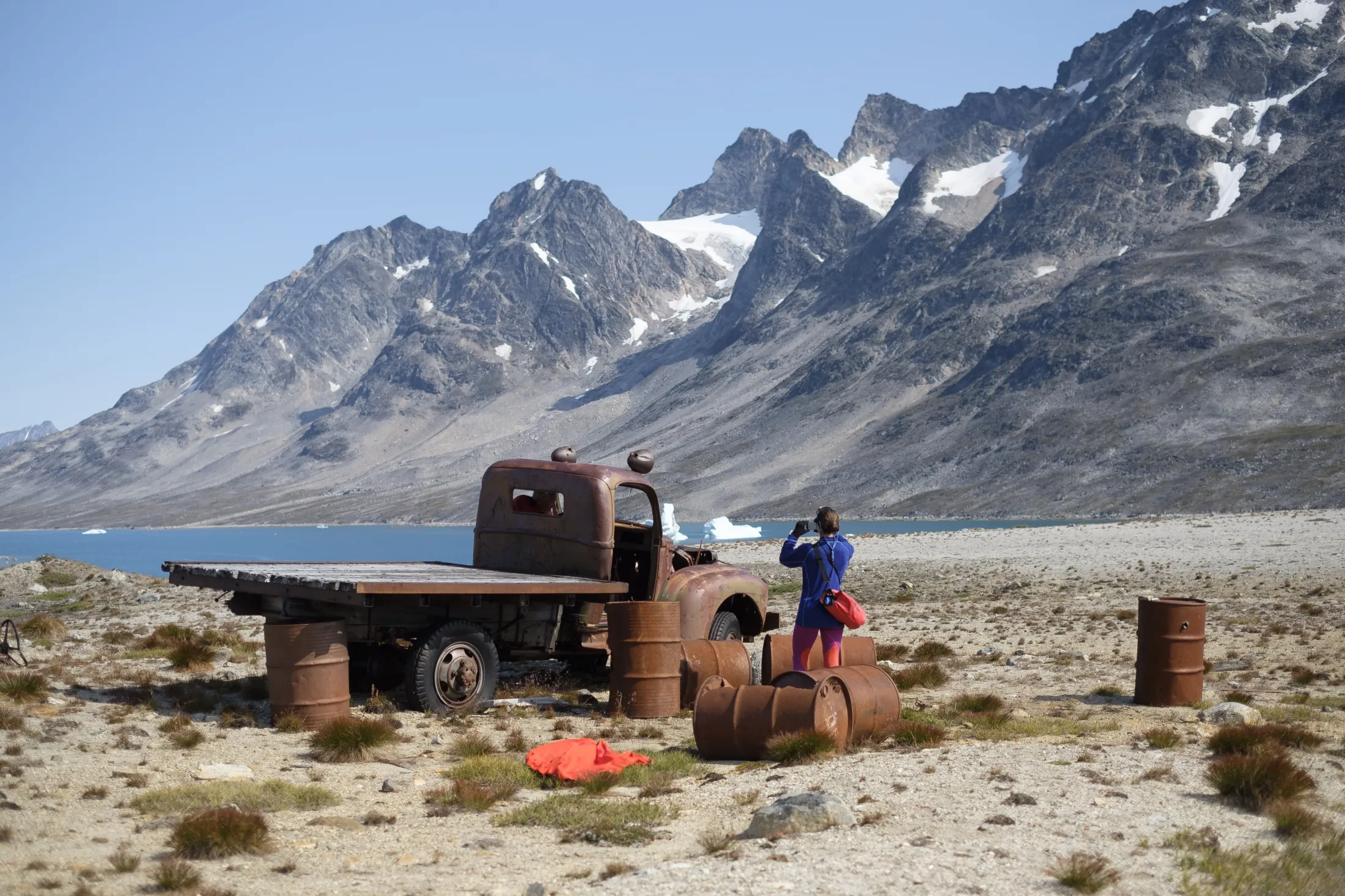 A person photographs an old, rusty truck wreck surrounded by rusty barrels on a plain, with a blue fjord and sharp, snow-dusted mountains in the background.