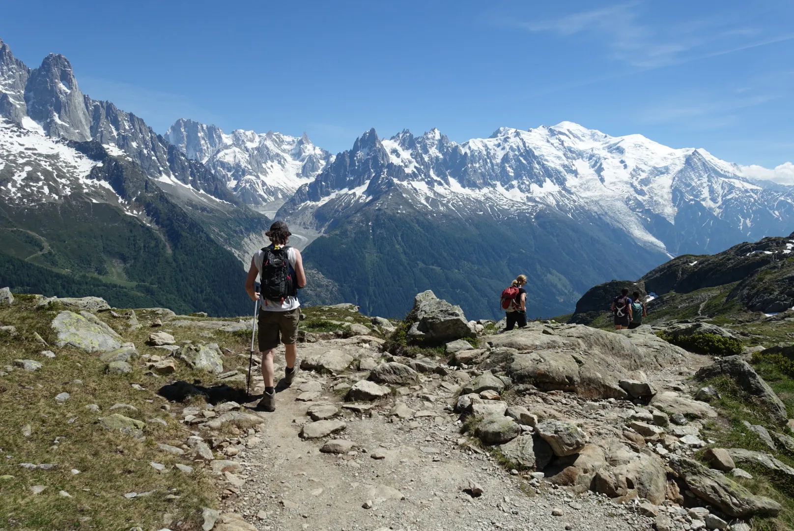 Hikers navigate a rocky high-mountain trail overlooking a massive valley and the snow-covered peaks of the Mont Blanc massif under a clear blue sky.