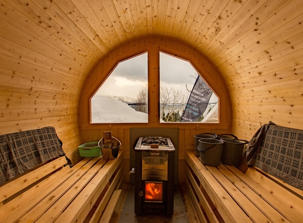 Interior of a wooden barrel sauna featuring benches, a wood-burning stove, and a window looking out at a snowy landscape.